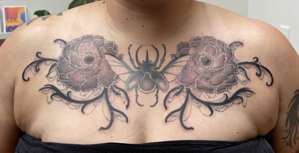 a tattoo of an atlas beetle in the center of the upper chest, flanked by a peony on either side, with black vining branches with cobwebs hanging off of them